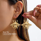 A close-up lifestyle shot of a model wearing gold bee drop earrings, with a hand holding one of the earrings to show detail. The model has brown hair and glittery pink nails, and the background is a clean white with text that reads 'Wear The Conversation Starter.