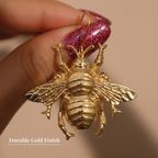 A gold bee-shaped earring with a textured finish, held by a hand with glittery pink nails against a warm beige background. A text overlay in the bottom-left corner reads 'Durable Gold Finish'.