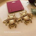 A pair of gold-tone bee-shaped drop earrings with detailed wings and legs, displayed on a beige surface with a magenta card behind them.