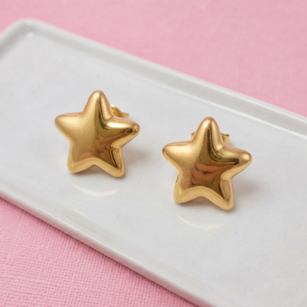 A pair of shiny gold star-shaped stud earrings resting on a white rectangular tray against a pink textured background.