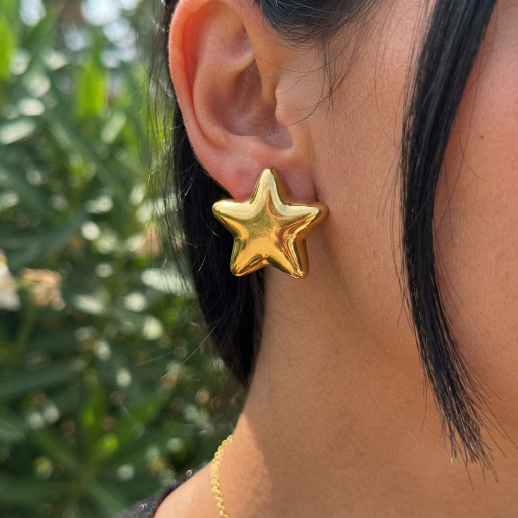 A close-up of a person's ear wearing a shiny gold star-shaped stud earring, with dark hair and a blurred green outdoor background.